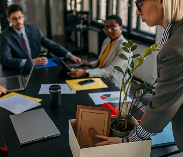 Employee packing personal items into a box while colleagues discuss in the background, representing voluntary redundancy in the workplace.